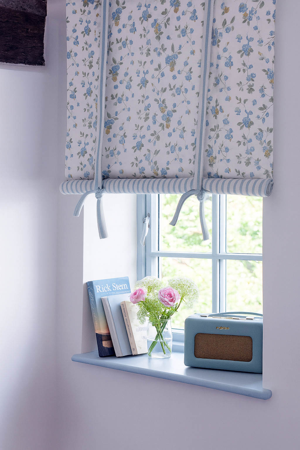 Estor Floral con Lazos en Azul y Blanco Estor con estampado floral azul sobre fondo blanco con lazos decorativos, en ventana pequeña con libros y flores